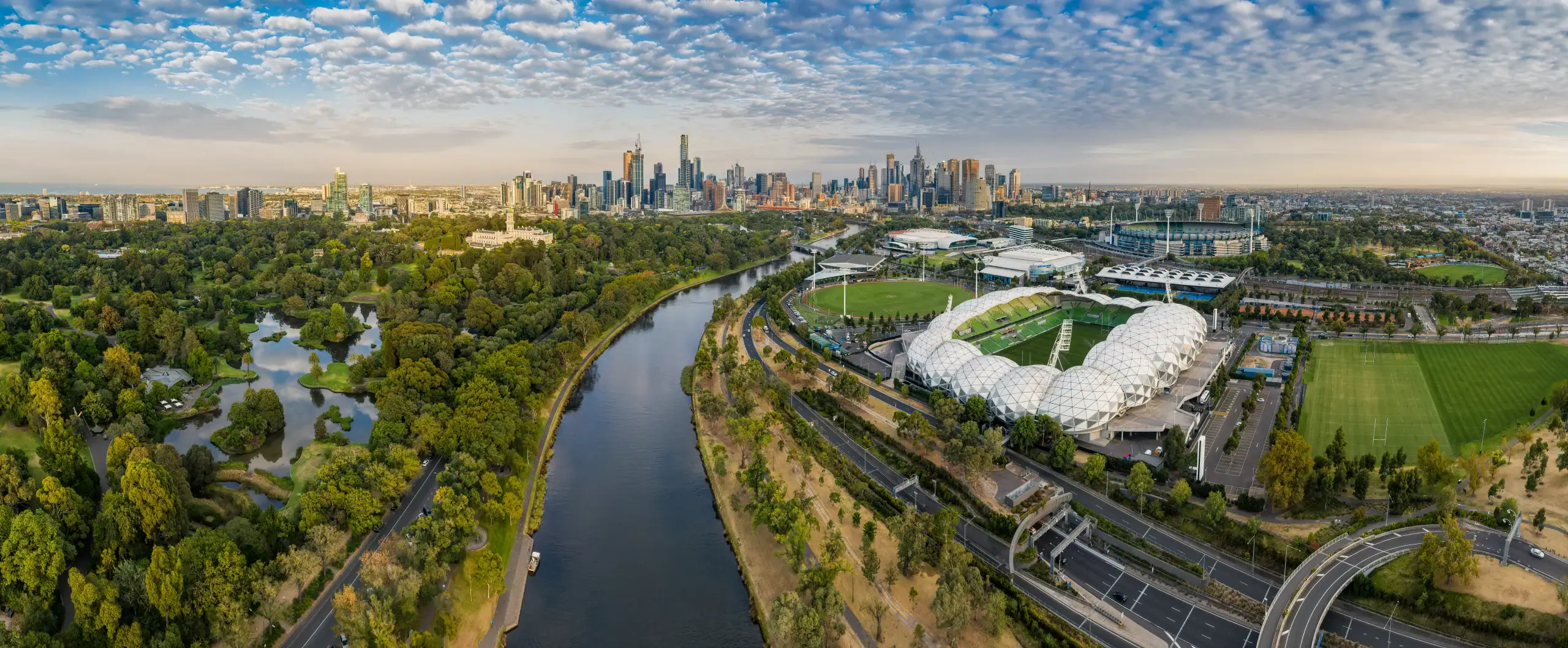 Aerial panoramic view of Melbourne CBD skyline with the Yarra River, Royal Botanic Gardens, AAMI Park, and Melbourne Park sports precinct under a cloudy sky